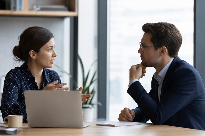 A professional woman explaining a work problem to her boss across a desk, pointing at a chart on a laptop.