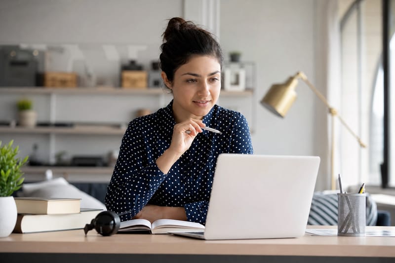 A student sits at a kitchen table in the morning, speaking aloud into a phone while a notebook and glass of water rest nearby.