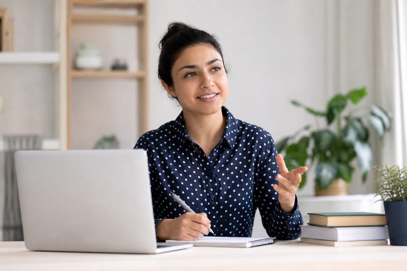 An examiner hands a small cue card to a candidate across a desk in a bright exam room.